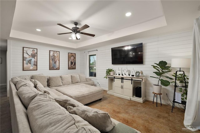 carpeted living room featuring a raised ceiling, wood walls, a ceiling fan, and recessed lighting