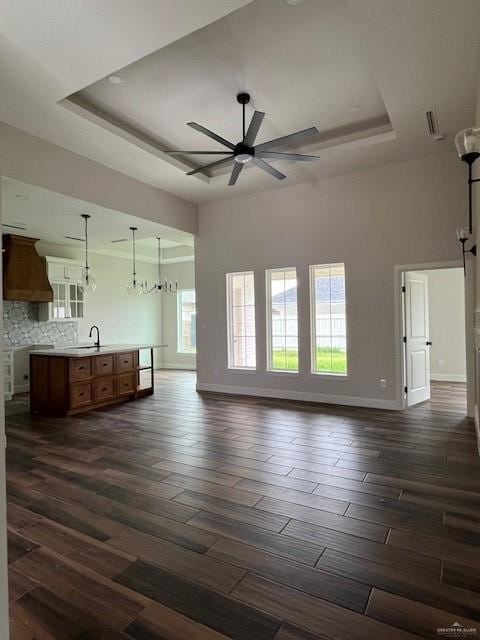 unfurnished living room featuring ceiling fan with notable chandelier, dark wood-type flooring, sink, and a raised ceiling