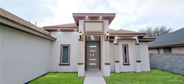 property entrance with a yard, roof with shingles, fence, and stucco siding