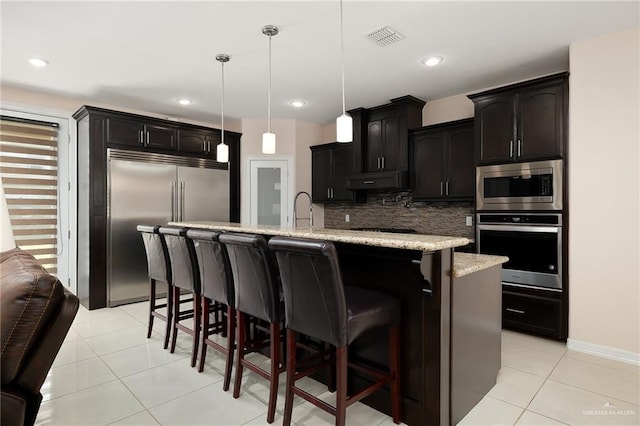 kitchen with decorative backsplash, light stone counters, a kitchen island with sink, built in appliances, and hanging light fixtures