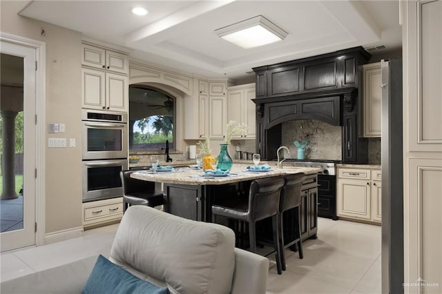 kitchen with a healthy amount of sunlight, a kitchen island with sink, cream cabinets, and stainless steel double oven