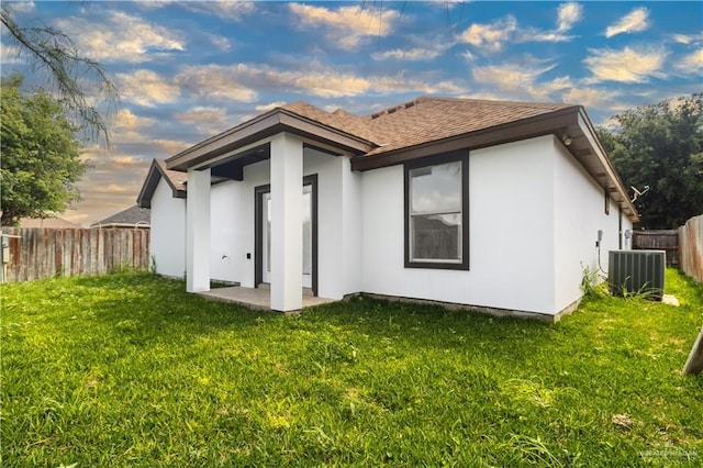 back of house with central AC, a fenced backyard, stucco siding, a shingled roof, and a yard