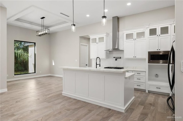 kitchen with white cabinetry, hanging light fixtures, stainless steel appliances, wall chimney range hood, and an island with sink