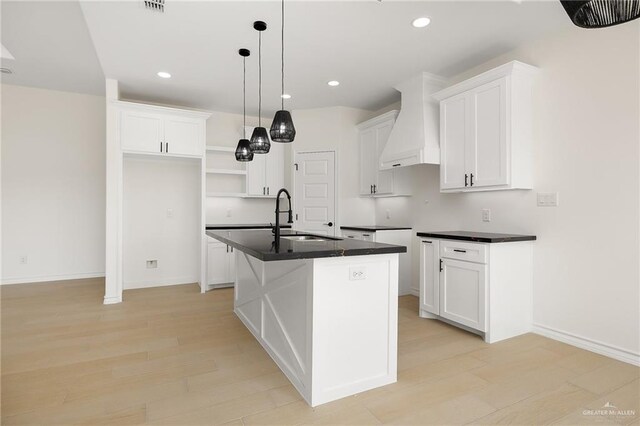 kitchen featuring decorative light fixtures, white cabinets, custom exhaust hood, a kitchen island with sink, and light wood-type flooring