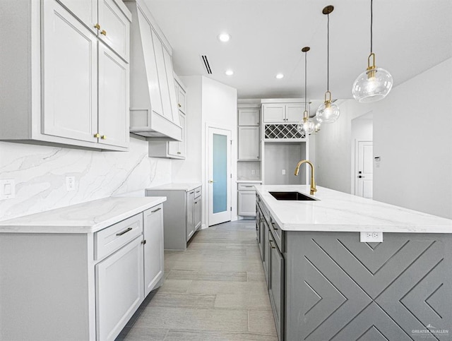 kitchen with a sink, gray cabinets, backsplash, light stone countertops, and a center island with sink