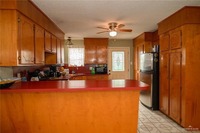 kitchen featuring a ceiling fan, a peninsula, freestanding refrigerator, a sink, and brown cabinets