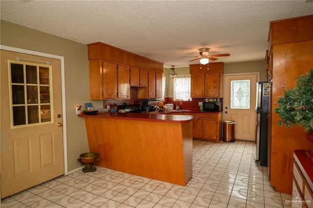 kitchen with a ceiling fan, brown cabinetry, a peninsula, freestanding refrigerator, and black microwave