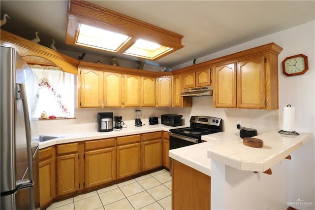 kitchen with tile counters, brown cabinetry, a peninsula, stainless steel appliances, and under cabinet range hood