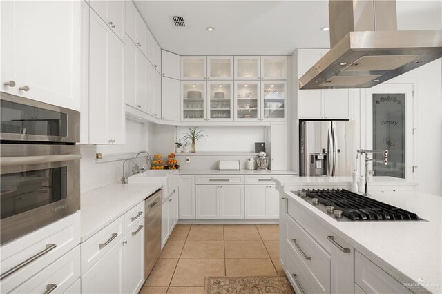 kitchen with wall chimney range hood, sink, light tile patterned floors, appliances with stainless steel finishes, and white cabinetry