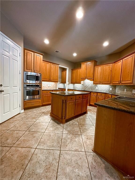 kitchen with light tile patterned flooring, dark stone counters, decorative backsplash, appliances with stainless steel finishes, and a kitchen island