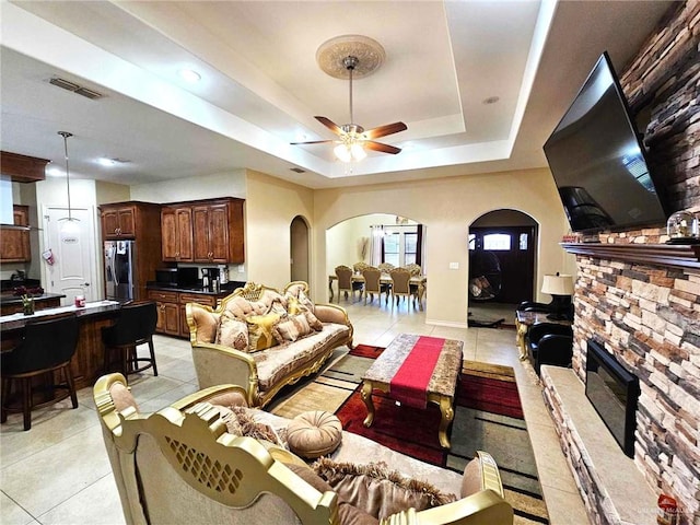 tiled living room featuring ceiling fan, a stone fireplace, and a tray ceiling