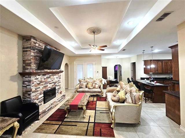 tiled living room with ceiling fan, a stone fireplace, a raised ceiling, and french doors