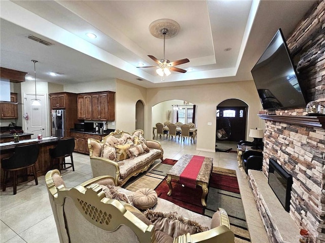 living room with a tray ceiling, a stone fireplace, ceiling fan, and light tile patterned floors