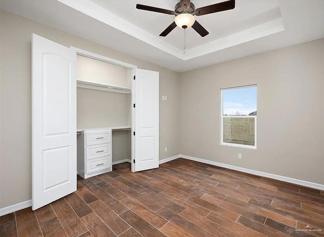 unfurnished bedroom with dark wood-style flooring, a closet, a raised ceiling, a ceiling fan, and baseboards