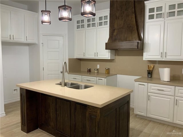 kitchen featuring custom exhaust hood, a kitchen island with sink, sink, decorative light fixtures, and white cabinets