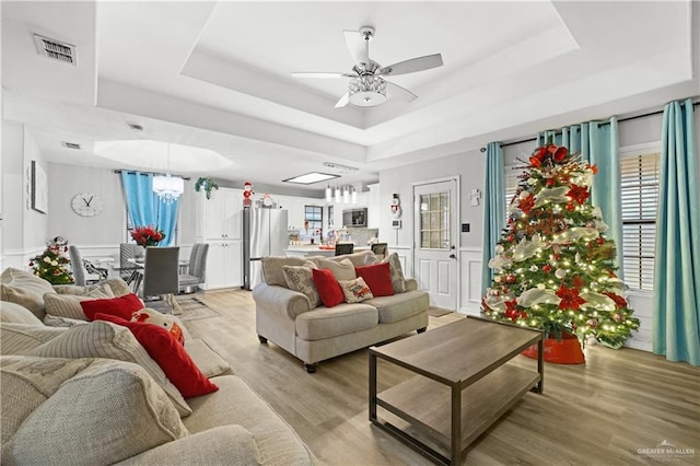 living room with a tray ceiling, ceiling fan with notable chandelier, and light wood-type flooring