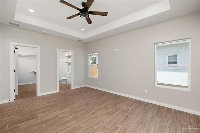 unfurnished bedroom featuring ensuite bathroom, a walk in closet, ceiling fan, light wood-type flooring, and a tray ceiling