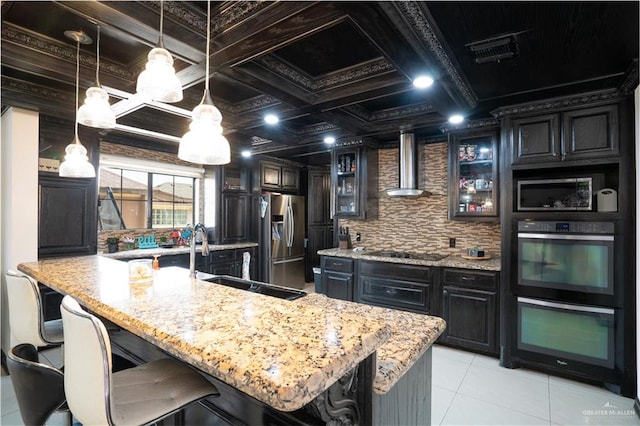 kitchen featuring coffered ceiling, black appliances, wall chimney range hood, hanging light fixtures, and a breakfast bar area