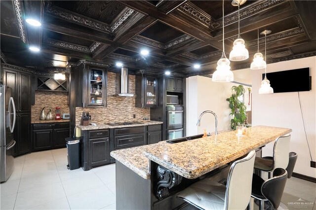 kitchen with sink, hanging light fixtures, wall chimney exhaust hood, an island with sink, and stainless steel appliances