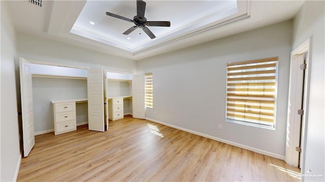 unfurnished bedroom with multiple closets, ceiling fan, a tray ceiling, and light hardwood / wood-style floors