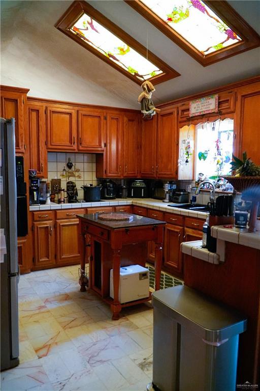 kitchen featuring tile countertops, brown cabinetry, decorative backsplash, a sink, and lofted ceiling