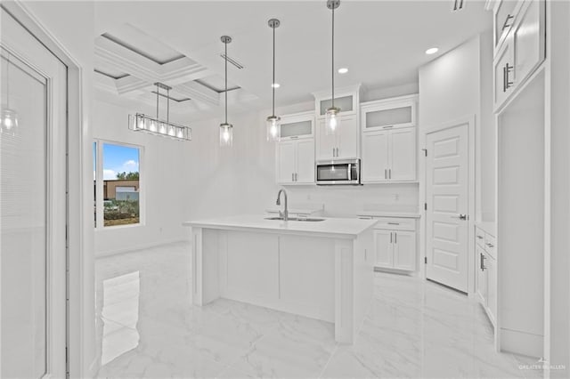 kitchen featuring light countertops, an island with sink, stainless steel microwave, glass insert cabinets, and pendant lighting