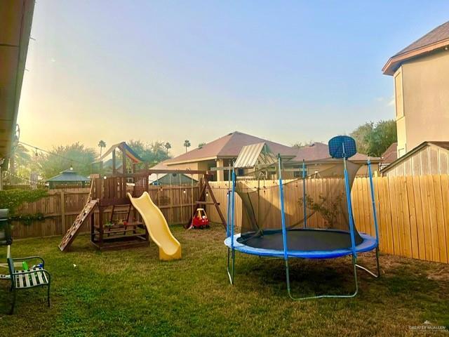 playground at dusk with a trampoline, a playground, a fenced backyard, and a lawn