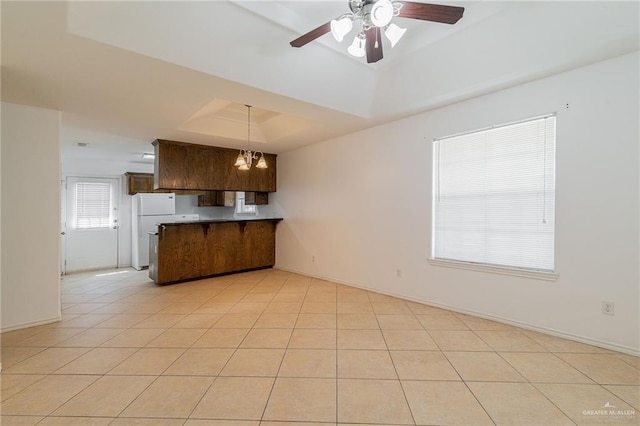 kitchen with light tile patterned floors, white refrigerator, a tray ceiling, ceiling fan with notable chandelier, and kitchen peninsula