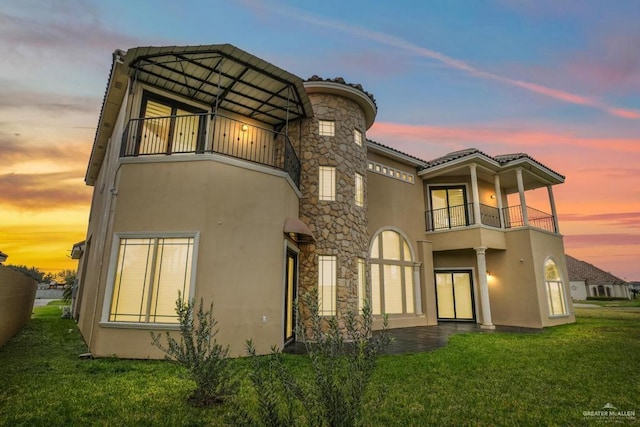 back house at dusk featuring a lawn and a balcony