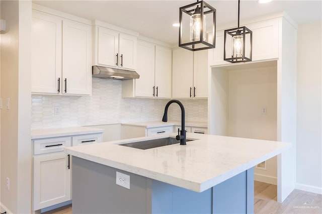 kitchen featuring a center island with sink, light stone countertops, under cabinet range hood, white cabinetry, and a sink