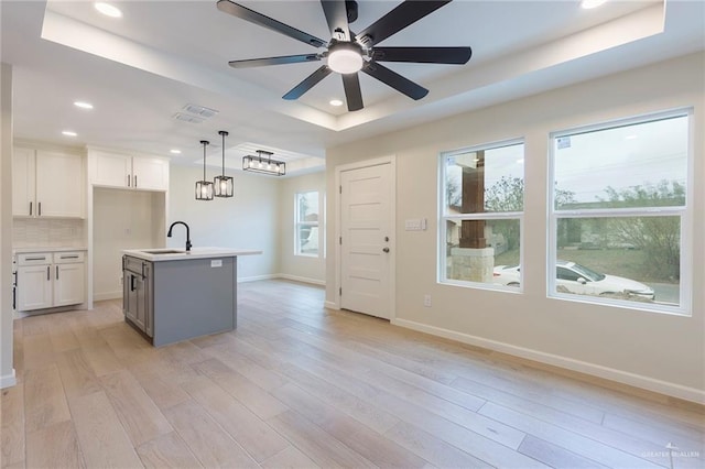 kitchen featuring a sink, light wood-style floors, decorative backsplash, a tray ceiling, and an island with sink