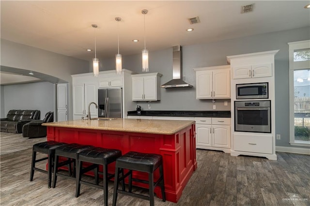 kitchen featuring wall chimney range hood, an island with sink, a kitchen bar, white cabinetry, and stainless steel appliances