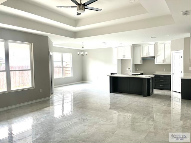 kitchen with ceiling fan with notable chandelier, visible vents, white cabinets, an island with sink, and a raised ceiling