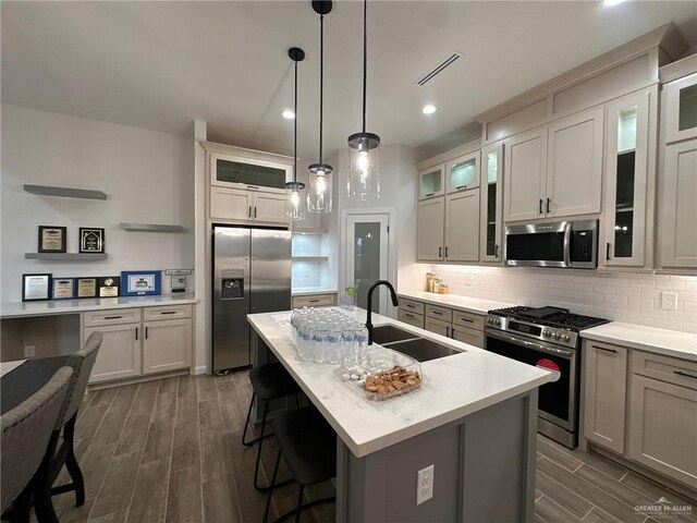 kitchen with decorative backsplash, appliances with stainless steel finishes, a kitchen island with sink, sink, and hanging light fixtures