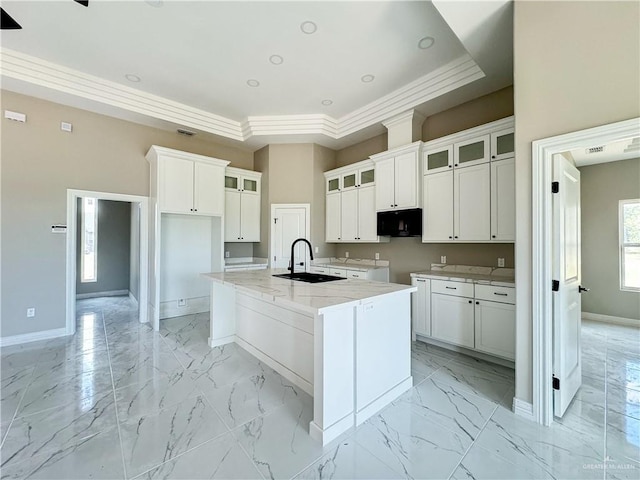 kitchen with light stone countertops, a raised ceiling, sink, white cabinets, and an island with sink