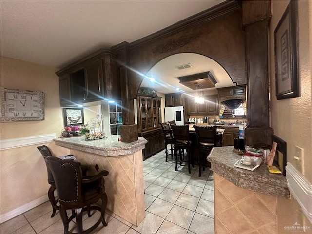 bar with dark brown cabinetry, white fridge with ice dispenser, light tile patterned floors, and ornamental molding