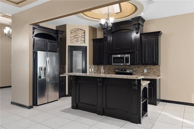 kitchen with an inviting chandelier, a tray ceiling, an island with sink, stainless steel appliances, and decorative backsplash