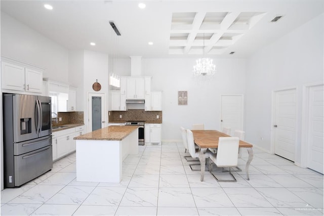 kitchen with appliances with stainless steel finishes, a center island, a towering ceiling, light stone counters, and coffered ceiling