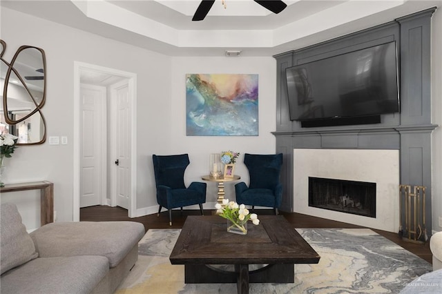 living room featuring a fireplace, wood finished floors, a ceiling fan, baseboards, and a tray ceiling