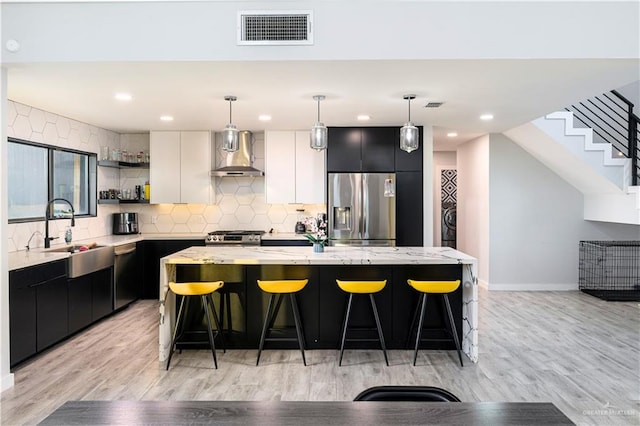 kitchen with stainless steel appliances, sink, wall chimney range hood, a kitchen island, and hanging light fixtures