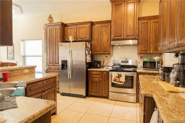 kitchen with light tile patterned floors, stainless steel appliances, brown cabinetry, and under cabinet range hood