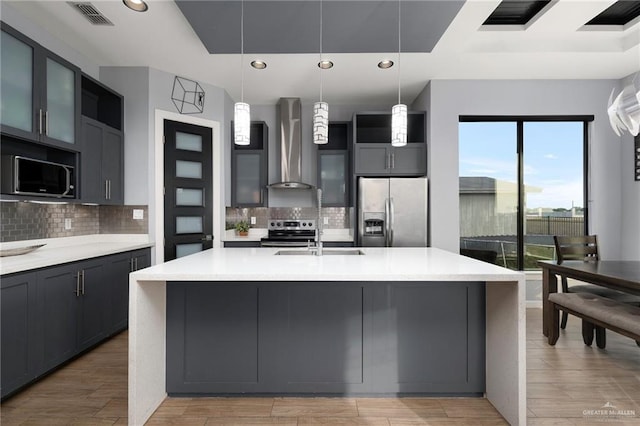 kitchen featuring wall chimney range hood, light hardwood / wood-style flooring, decorative backsplash, an island with sink, and stainless steel appliances