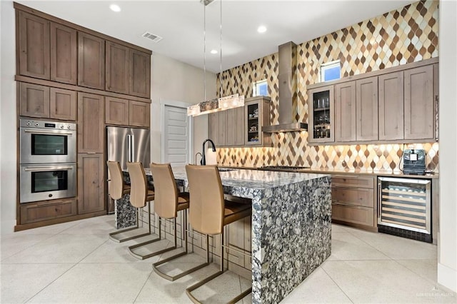 kitchen with wine cooler, a center island with sink, hanging light fixtures, and wall chimney range hood