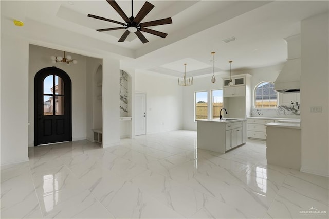 kitchen with a tray ceiling, marble finish floor, light countertops, hanging light fixtures, and an island with sink