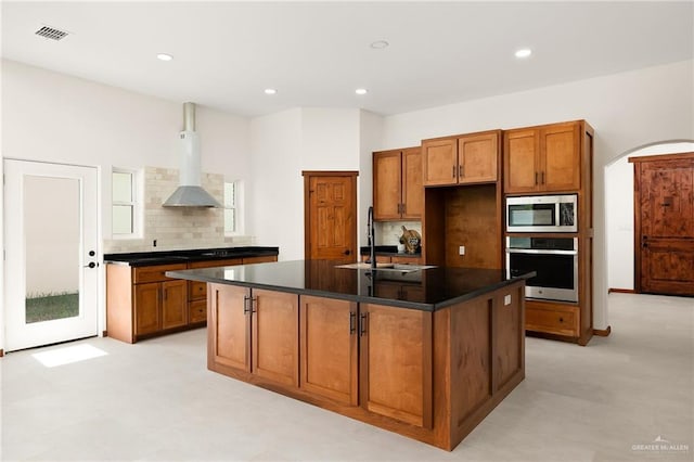 kitchen featuring sink, stainless steel appliances, wall chimney range hood, backsplash, and a center island with sink