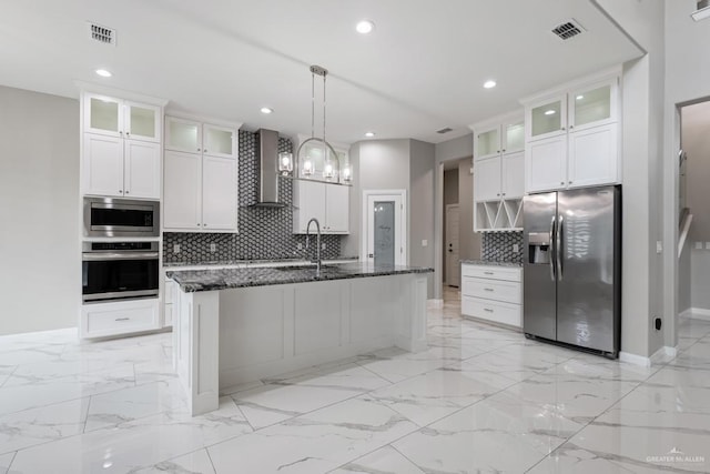kitchen with appliances with stainless steel finishes, an island with sink, dark stone counters, wall chimney range hood, and white cabinetry
