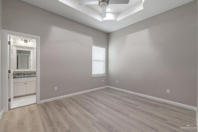 unfurnished bedroom with light wood-type flooring, connected bathroom, a tray ceiling, sink, and ceiling fan