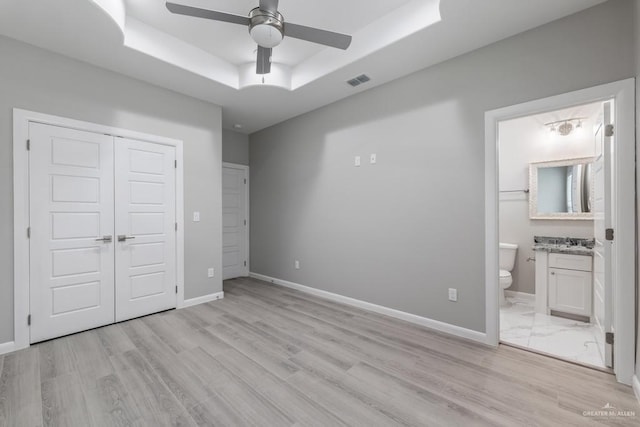 unfurnished bedroom with a closet, light hardwood / wood-style flooring, a tray ceiling, connected bathroom, and ceiling fan