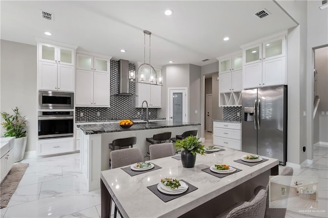 kitchen featuring wall chimney range hood, an island with sink, stainless steel appliances, and a breakfast bar area