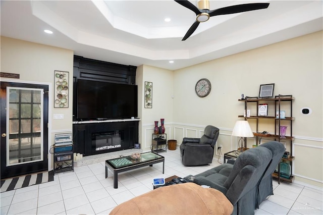 living area with light tile patterned floors, a raised ceiling, a ceiling fan, wainscoting, and a decorative wall